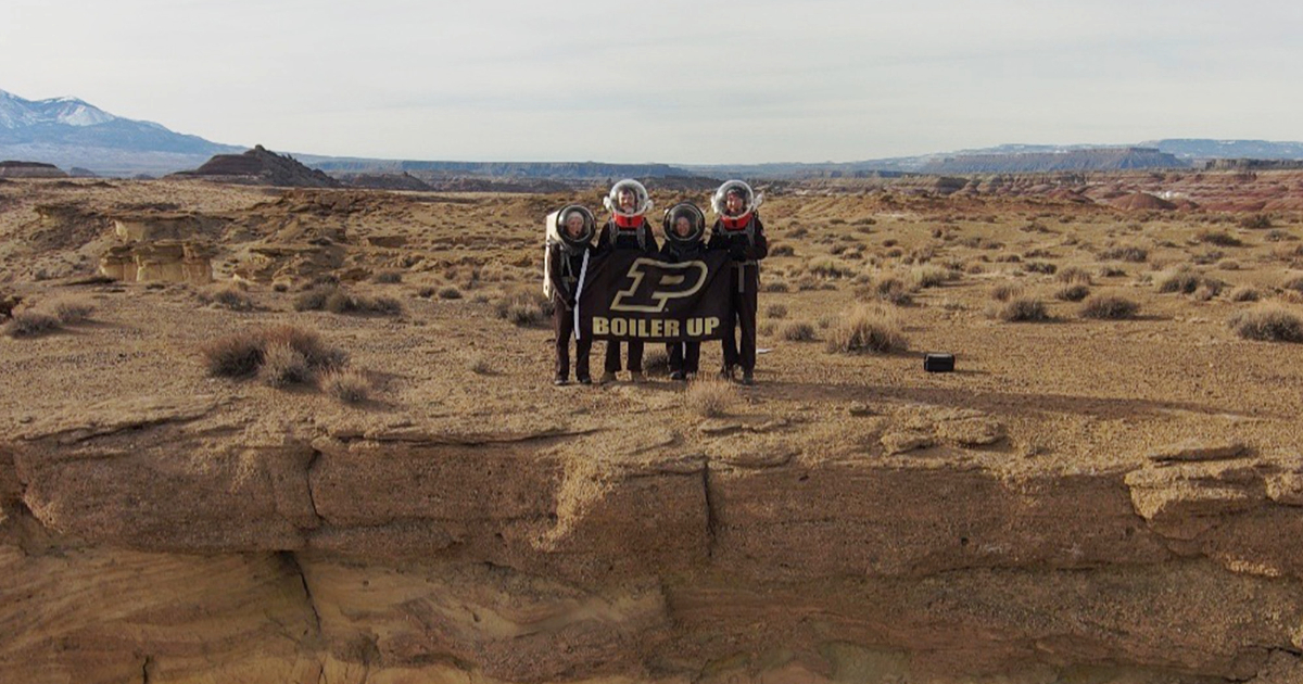 Students holding a Purdue flag and standing in a desert, wearing simulated Mars spacesuits