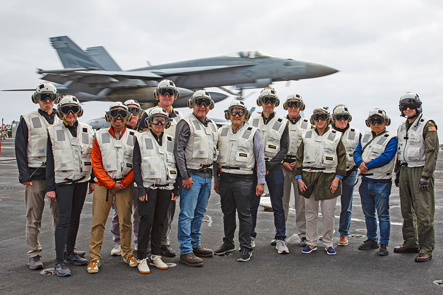 A group of people with goggles and hearing protection standing on the flight deck of an aircraft carrier as a fighter jet takes off in the background