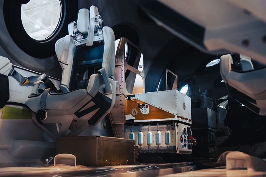 two metal boxes holding automated experiments are shown mounted behind a passenger seat inside the VSS Unity spacecraft