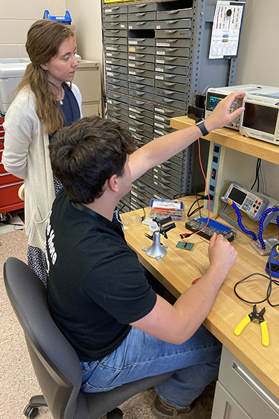 A student at a desk is demonstrating lab equipment to an instructor who stands behind.