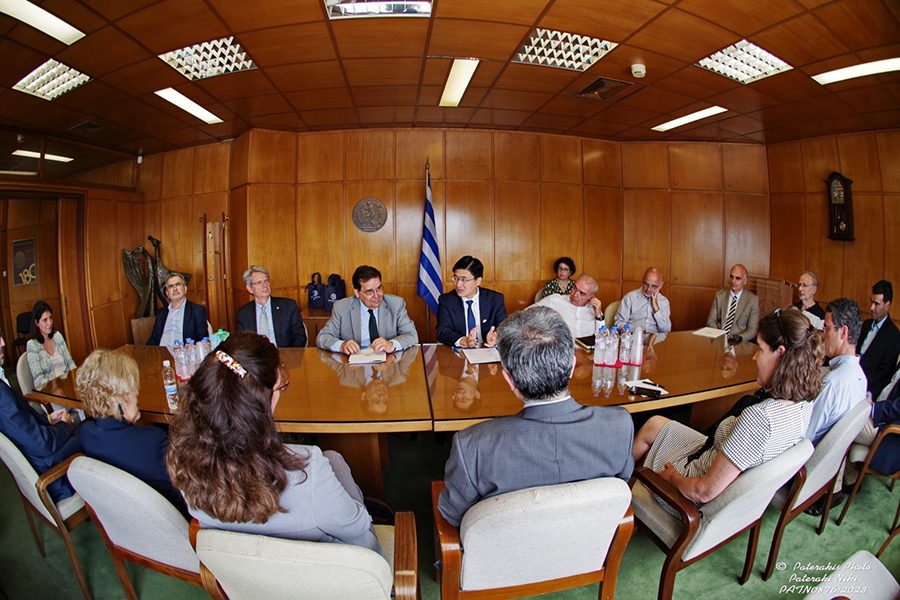 Representatives from various Greek universities and organizations and Purdue University are gathered around a long table. Behind them is a Greek flag.