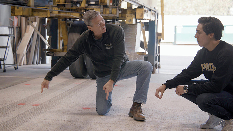 Professor John Haddock and graduate student Oscar Moncada examine a slab of concrete pavement they are testing to handle heavy truck loads with wireless power transfer technology installed below the surface. The machine behind them is designed to imitate those loads by repeatedly passing half a loaded semi-truck axle across the concrete slab.