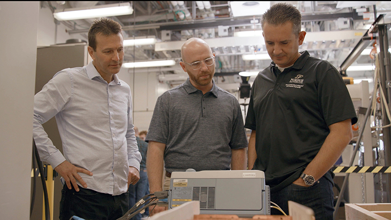 In a Purdue electrical engineering lab, Steve Pekarek, from left, Aaron Brovont and Dionysios Aliprantis stand before a gray box which is an instrument that will measure the electromagnetic performance of coils they are developing to transmit power to receiver coils on electric vehicles as they drive.