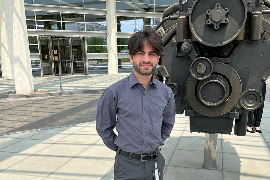 Hasan, dressed in dark slacks and a button up shirt, stands in front of an antique engine at the Cummins corporate office.