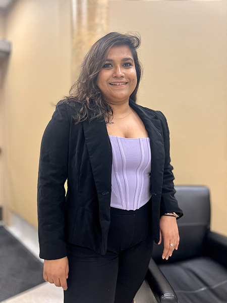 dressed in a black suit with a fitted lilac top, Somosmita Mitra stands beside a conference table