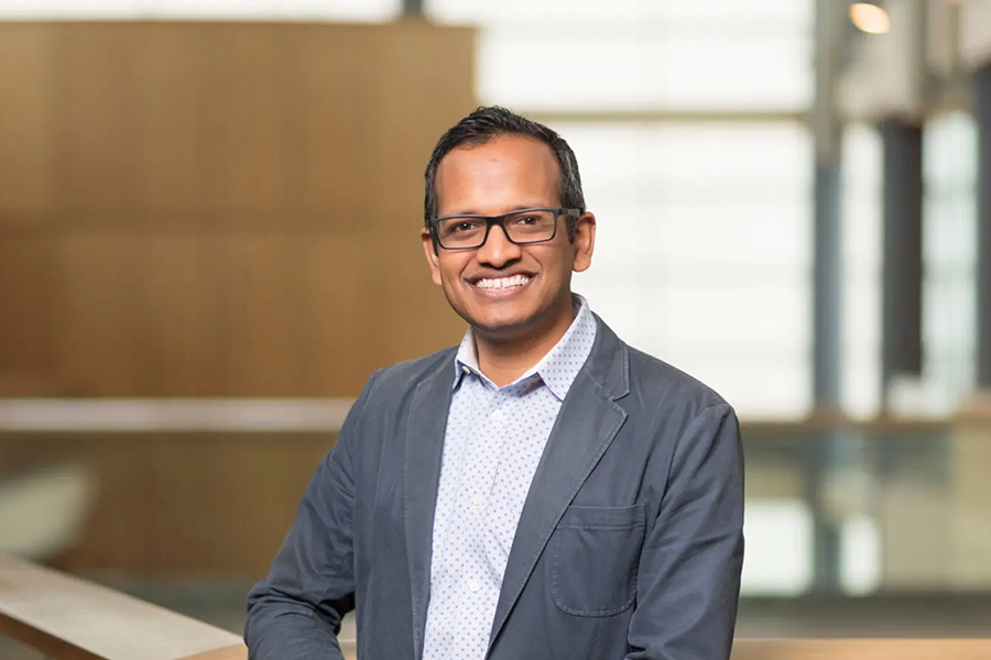 Portrait of Milind Kulkarni standing in front of a railing on the second story of a building. He is wearing a button up shirt, suit jacket, and glasses.