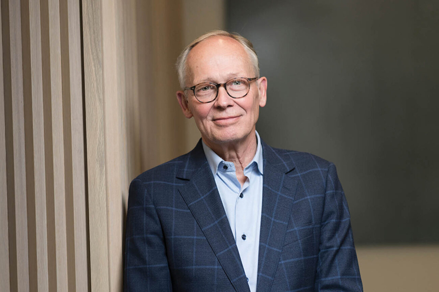 Portrait of Mark Lundstrom. He is wearing a blue suit with a checkered pattern and is leaning against a wall.