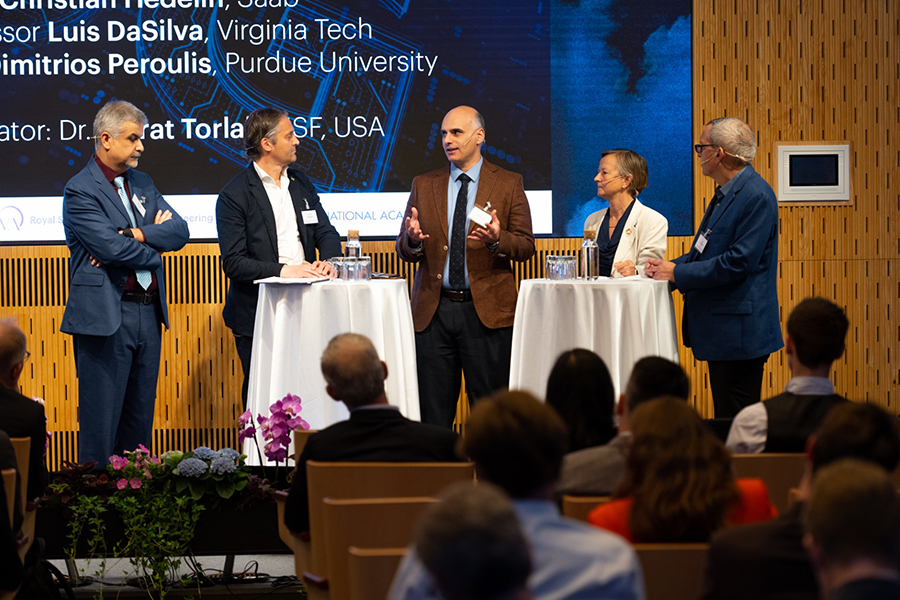 Professor Dimitri Peroulis stands in the center of other panel members. They are on stage with an audience in the foreground.