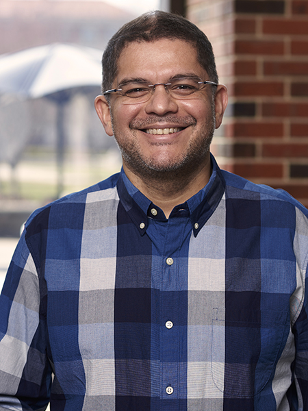 Portrait of Sunil Bhave. He is wearing a blue and white plaid shirt and standing in front of a window.