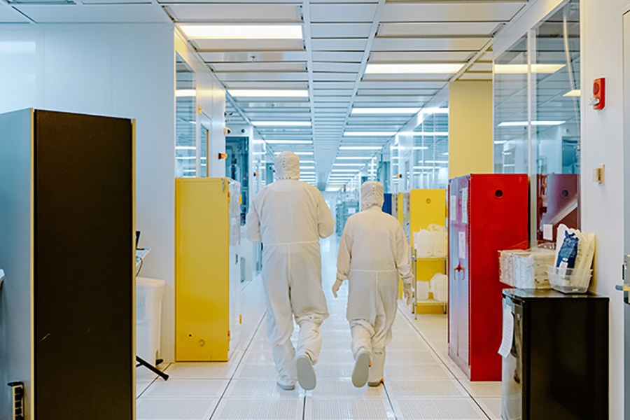 Two cleanroom workers are walking away from the camera wearing full white coveralls.