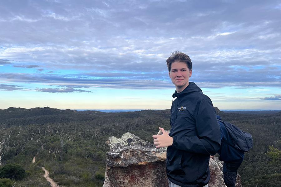 Grady Arnold poses for a photo by a rock while hiking. In the distance you can see a sandy forest terrain and the ocean.