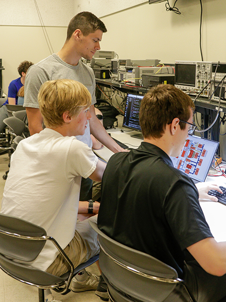 Three students are working at a desk in an electrical and computer engineering lab. One student has control of the keyboard while the other two are looking at a screen that displays information.