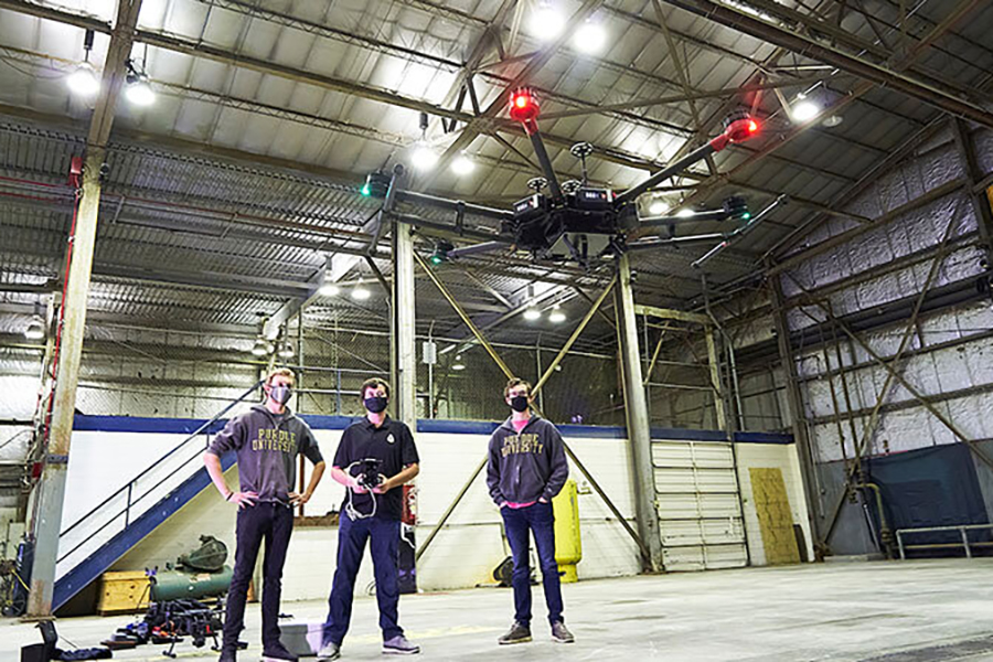 3 males students, wearing Purdue apparel, stand in a large building. The middle student holds the controls for a done that is flying in the foreground.