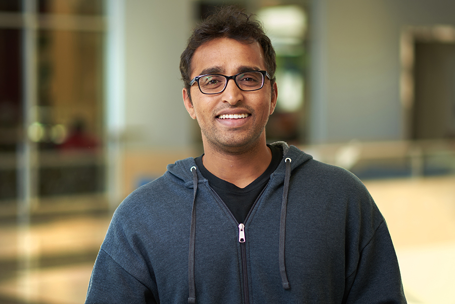 A smiling young man wearing glasses and a gray zip-up jacket, poses in front of a blurred background