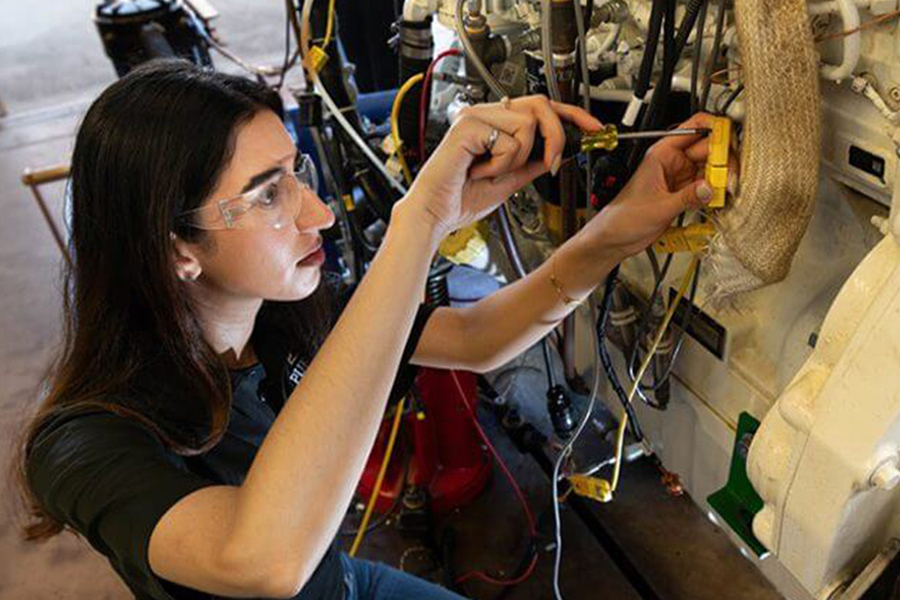 A woman, wearing safety glasses, uses a screwdriver to work on an AI-connected piece of equipment.