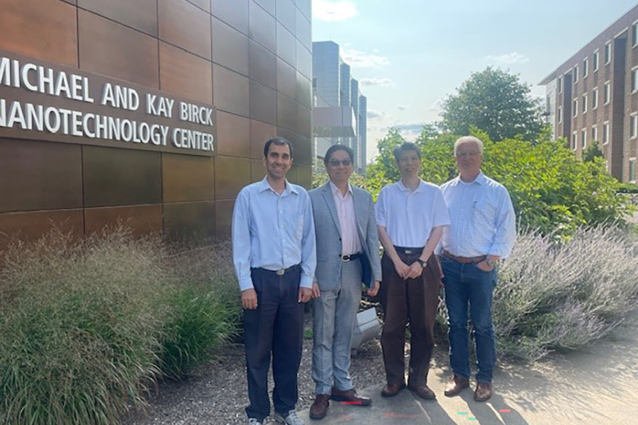 Four men stand side-by-side posing in front of a brick building on a sunny day