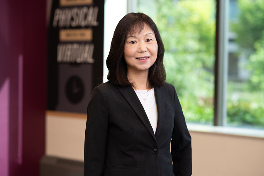 Dan Jiao is posing for a portrait in the Rice Design Studio. Behind her is a set of windows and a wall mural. She is wearing a white top with a black jacket.