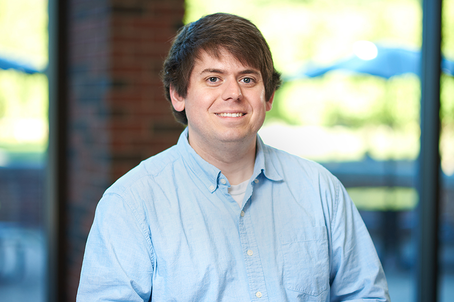A man wearing a blue shirt stands confidently in front of a textured brick wall
