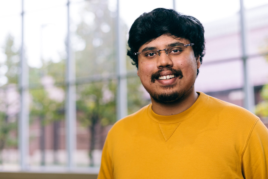 Professor Makur poses for a portrait in the atrium of the CS building. He is wearing glasses and an orange t shirt.