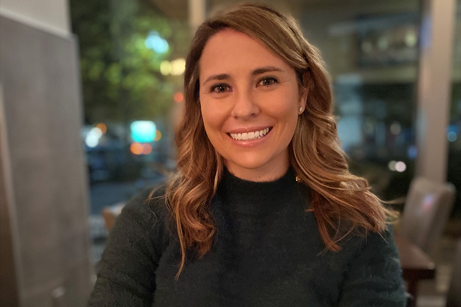 Portrait of Julia Filiberti Allen. She is seated at a table, wearing a sweater, with windows in the background.