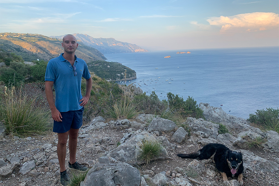 Giorgio, wearing blue shorts and a blue button up shirt, poses for a portrait with the Amalfi Cost in the background. To the viewer's right, there is a black dog also in the picture.