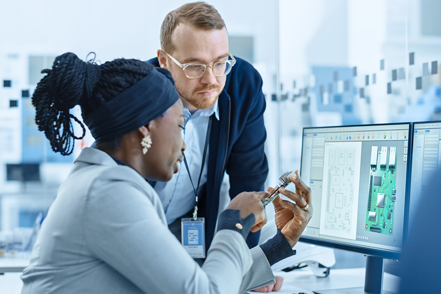 In this photo, a woman is sitting at a desk holding a piece of cuircuitboard. She is sitting in front of a computer. A man stands to her side examining the chip.
