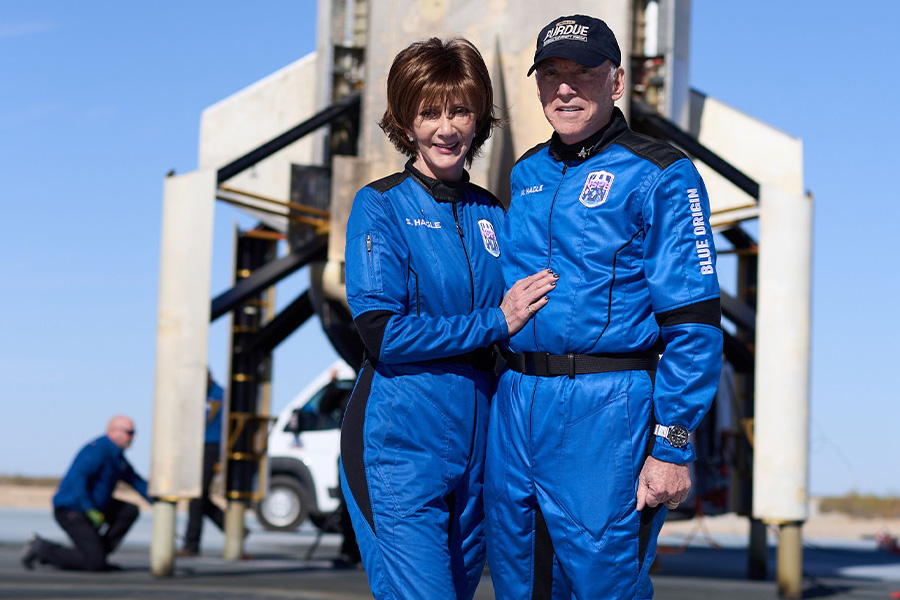 A man and woman in blue astronaut suits stand smiling in front of a rocket. The sunny sky is clear. Another person works in the background.