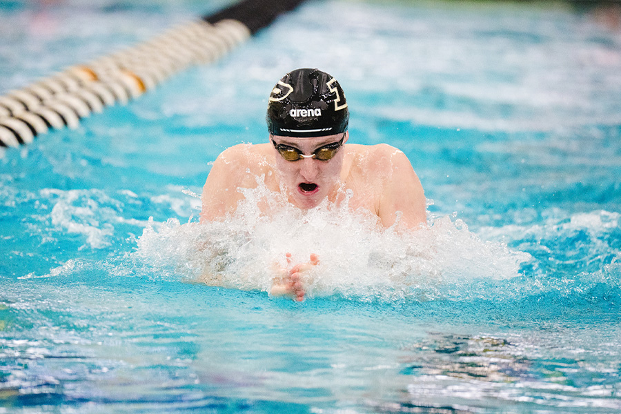 A swimmer wearing goggles and a black swim cap competes in a pool, performing the breaststroke. Water splashes outward, conveying focus and motion.