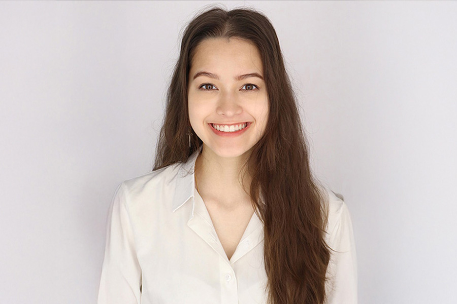 A woman with long brown hair smiles warmly, wearing a white blouse. She stands against a plain background, conveying a friendly and approachable tone.