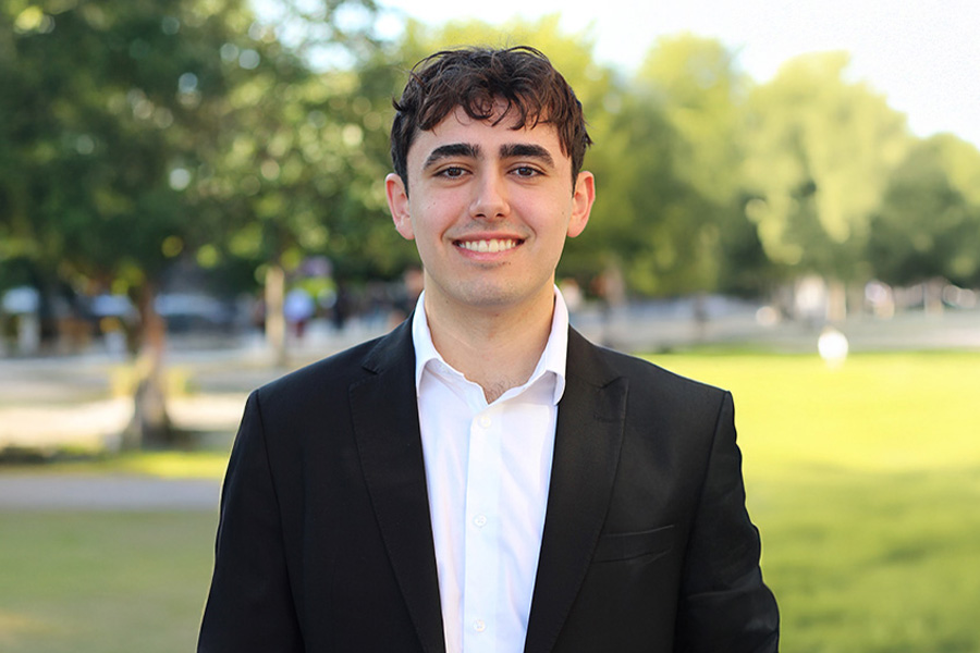 Smiling young man in a black suit and white shirt stands outdoors in a park. Bright, sunny day with green trees providing a serene backdrop.