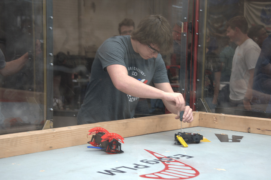 A person in a gray shirt adjusts a small battle robot inside an arena with a screwdriver. Another red robot is nearby. Onlookers watch through glass.