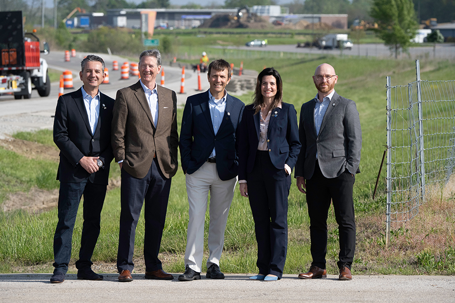 Five people in suits stand smiling on a roadside near a construction site with traffic cones and vehicles, conveying a professional and positive tone.