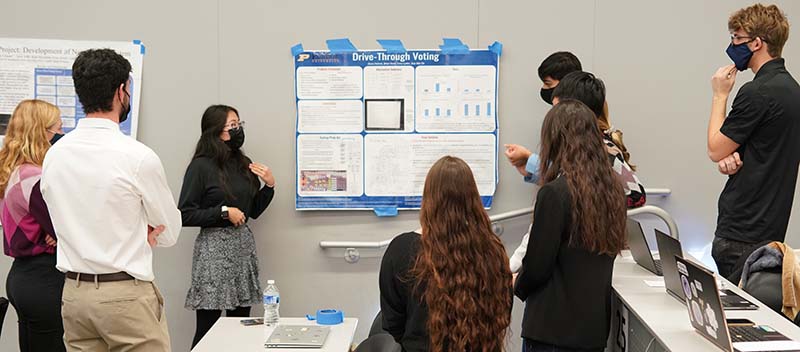 Students gather around a poster that exhibits an engineering design project for voting technology.